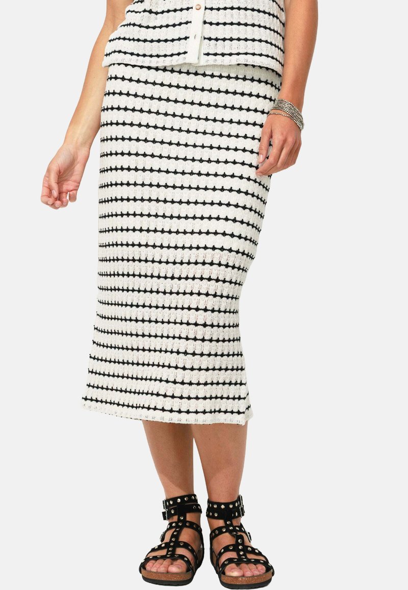 Jupe tricotée en blanc avec des rayures horizontales noires, présentant une coupe ajustée. Associée à des sandales noires à lanières avec des accents cloutés.