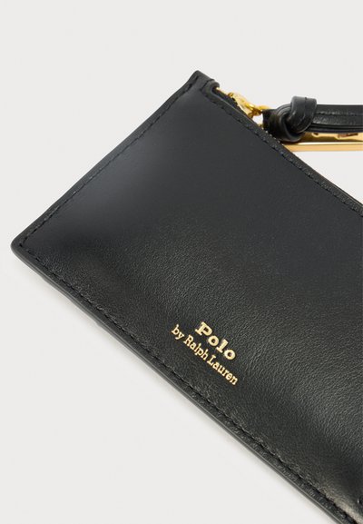 Crna kožna novčanica sa zlatnim detaljima logotipa. Ima glatku teksturu, ukrase od šavova i priloženi remen za jednostavno rukovanje.