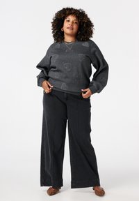 Vrouw met krullend haar, gekleed in een donkergrijze trui met hartpatronen, een brede broek in het zwart en flatjes met luipaardprint, staand tegen een witte achtergrond.