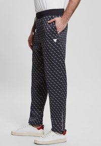 Muž v černých vzorovaných teplácích s bílým potiskem loga a bílých teniskách s červeným detailem na patě, stojící s jednou rukou v kapse.