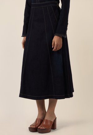 Frau trägt einen dunkelblauen Denim-Midirock mit A-Linien-Schnitt und braune offene Plateau-Pumps, steht vor einem schlichten hellen Hintergrund.