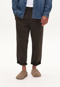 Dunkelbraune, locker sitzende Hosen mit umgeschlagenen Bündchen, kombiniert mit beigen Slippers. Dazu wird ein Jeanshemd mit Knopfleiste getragen.