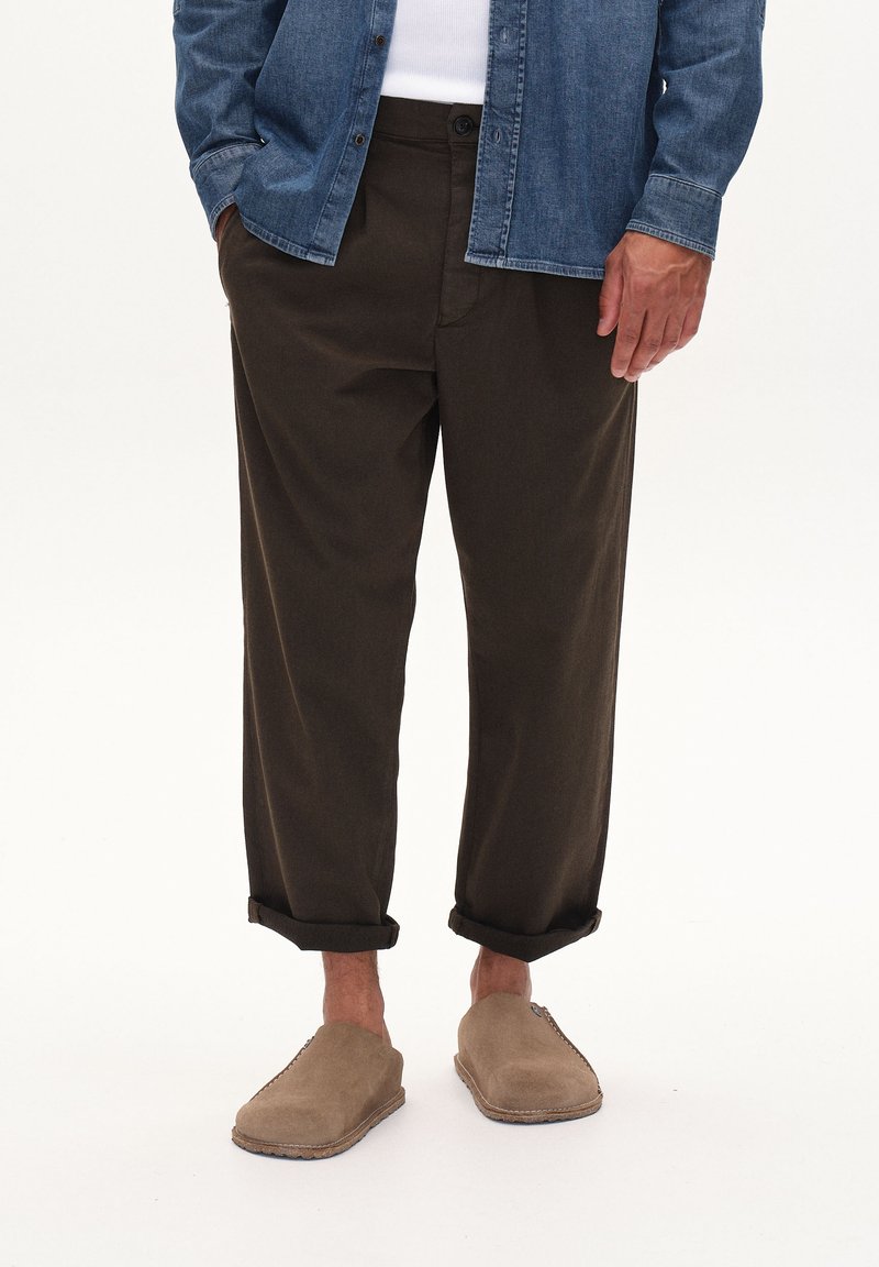 Dunkelbraune, locker sitzende Hosen mit umgeschlagenen Bündchen, kombiniert mit beigen Slippers. Dazu wird ein Jeanshemd mit Knopfleiste getragen.