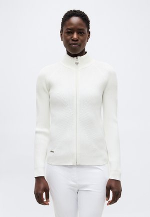 Femeie purtând un pulover alb strâmt cu fermoar, cu model cu dungi și guler înalt, și pantaloni albi, stând în fața unui fundal simplu, deschis la culoare.