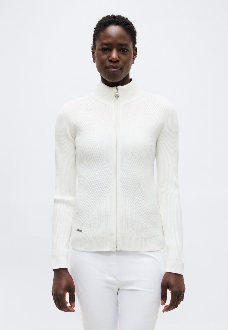 Γυναίκα που φοράει ένα εφαρμοστό λευκό ριμπ πλεκτό πουλόβερ με φερμουάρ και ψηλό γιακά και λευκό παντελόνι, στέκεται μπροστά σε απλό ανοιχτό φόντο.