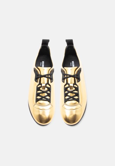Pár lesklých zlatých tenisiek s čiernymi šnúrkami a vnútrom, zobrazených zhora na bielom pozadí.