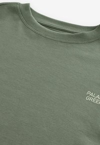 Gros plan d'un sweat-shirt vert à col rond avec col côtelé et texte blanc partiel indiquant "PALACE GREEN" sur la poitrine.
