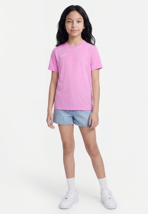 Fille aux longs cheveux foncés portant un t-shirt rose Nike, un short en jean bleu clair, des chaussettes blanches et des baskets blanches, debout devant un fond blanc.