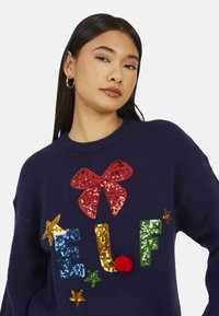 Maglione blu navy con un grande fiocco rosso di paillettes e lettere colorate di paillettes che formano la parola "ELF", decorate con stelle e un pon pon.