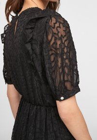 Svart klänning med genomskinliga, texturerade ärmar med broderade blommönster. Volangdetaljer och knäppning bak.