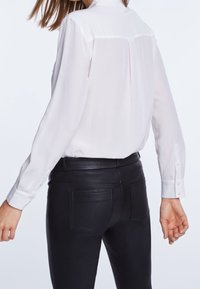 Femme vêtue d'un chemisier blanc transparent à manches longues, rentré dans un pantalon en cuir noir à taille haute, vue de dos.