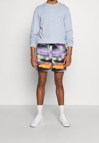 Světle modrý top s dlouhým rukávem zkombinovaný s šortkami s barevným tie-dye vzorem v purpurové, oranžové a černé barvě. Bílé tenisky nošené s bílými ponožkami.