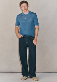 Blaues kurzärmeliges Polo mit strukturierter Strickoberfläche, Drei-Knopfleiste, kombiniert mit lockeren, dunklen Jeans und hellen Schuhen vor neutralem Hintergrund.