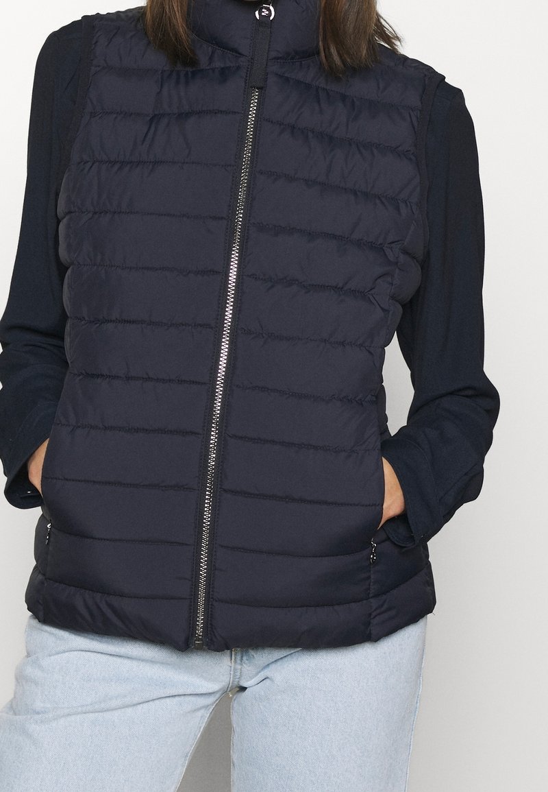 Chaleco acolchado azul marino con cuello alto, cremallera frontal y bolsillos laterales. Presenta un patrón de costuras horizontales y una textura suave.