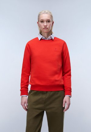 Kvinde med kort blondt hår iført en rød sweatshirt over en stribet skjorte med krave og olivengrønne bukser mod en ensfarvet lys baggrund.