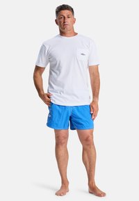 White cotton t-shirt with a chest pocket, paired with blue swim shorts. The shorts feature a logo and a smooth texture. Model stands barefoot.