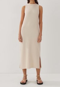 Robe côtelée sans manches couleur beige clair, avec fentes sur les côtés, longueur au genou et col rond. Texture douce et silhouette ajustée.