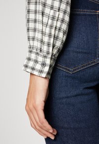 Chemise à carreaux blanche et noire à boutons, en tissu doux, avec des manches retroussées, associée à un jean en denim bleu foncé.