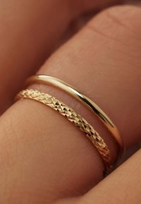 Deux anneaux en or sur un doigt : l'un lisse et poli, l'autre texturé avec des motifs géométriques, mettant en valeur un contraste dans le design et la finition.