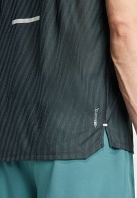 Schwarzes Sportshirt mit vertikalen Strukturmustern, einem seitlichen Schlitz und einem weißen Akzentstreifen auf der Brust. Der Stoff ist leicht und atmungsaktiv.