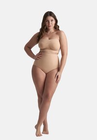 Donna che indossa un body senza spalline color carne con slip a vita alta, in piedi scalza con una mano sui fianchi su uno sfondo bianco.