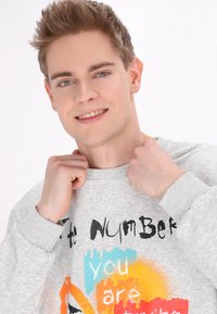 Hanorac gri cu gât rotund, având text negru și stropi colorati de portocaliu și albastru. Manșetele și tivul cu textură ribbată adaugă un plus de stil.
