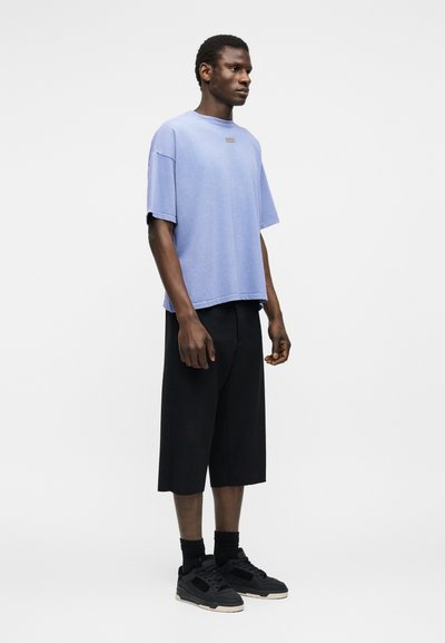 Άνδρας που στέκεται φορώντας ένα ανοιχτό μωβ φαρδύ κοντομάνικο πουκάμισο, μαύρες φαρδιές βερμούδες μέχρι το γόνατο, μαύρες κάλτσες και μαύρα αθλητικά παπούτσια μπροστά σε λευκό φόντο.