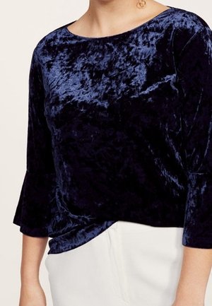 Femme portant un top en velours froissé bleu foncé à manches trois-quarts et un pantalon blanc.