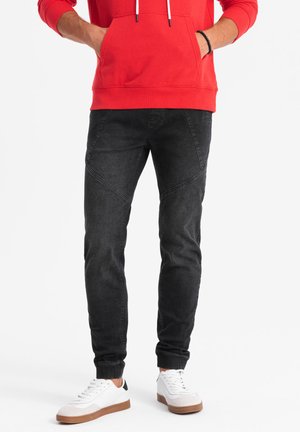 Pantalones vaqueros negros con un corte ajustado, que presentan costuras angulares y una textura de denim suave. Combinados con una sudadera roja con capucha y zapatillas blancas.