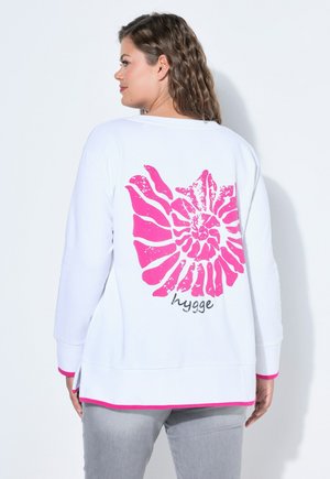 Frau trägt weißen Pullover mit leuchtend pinkem abstraktem Muscheldesign und dem Schriftzug "hygge" auf dem Rücken, kombiniert mit hellgrauen Hosen.