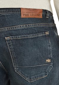 Tamno plave traperice s teksturiranim završetkom. Imaju smeđu kožnu oznaku s natpisom "PME LEGEND" i malim logotipom na stražnjem džepu.