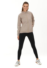 Ljus beige zip-up jacka med hög krage och reflexdetaljer, i kombination med svarta leggings och silverfärgade träningsskor.