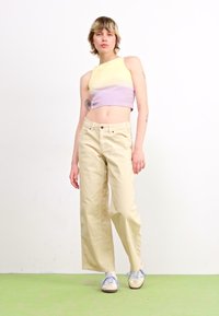Un crop top côtelé lilas et jaune, un pantalon large taille haute beige, assortis à des baskets bleu pastel sur un sol vert.