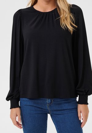 Femme portant un chemisier noir à manches longues avec poignets et encolure froncés, associée à un jean en denim bleu, debout devant un fond uni.