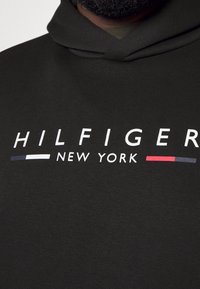 Felpa nera con cappuccio e un grande logo bianco che recita "HILFIGER NEW YORK". Presenta strisce orizzontali blu e rosse come accenti sotto il testo.
