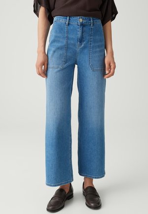 Femme portant un jean bleu taille haute à jambes larges avec des poches plaquées à l’avant et des mocassins marron foncé, debout devant un fond clair uni.