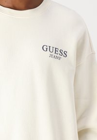 Felpa color crema realizzata in morbido tessuto, con collo a giro e un piccolo logo "GUESS JEANS" in navy sul lato sinistro del petto.