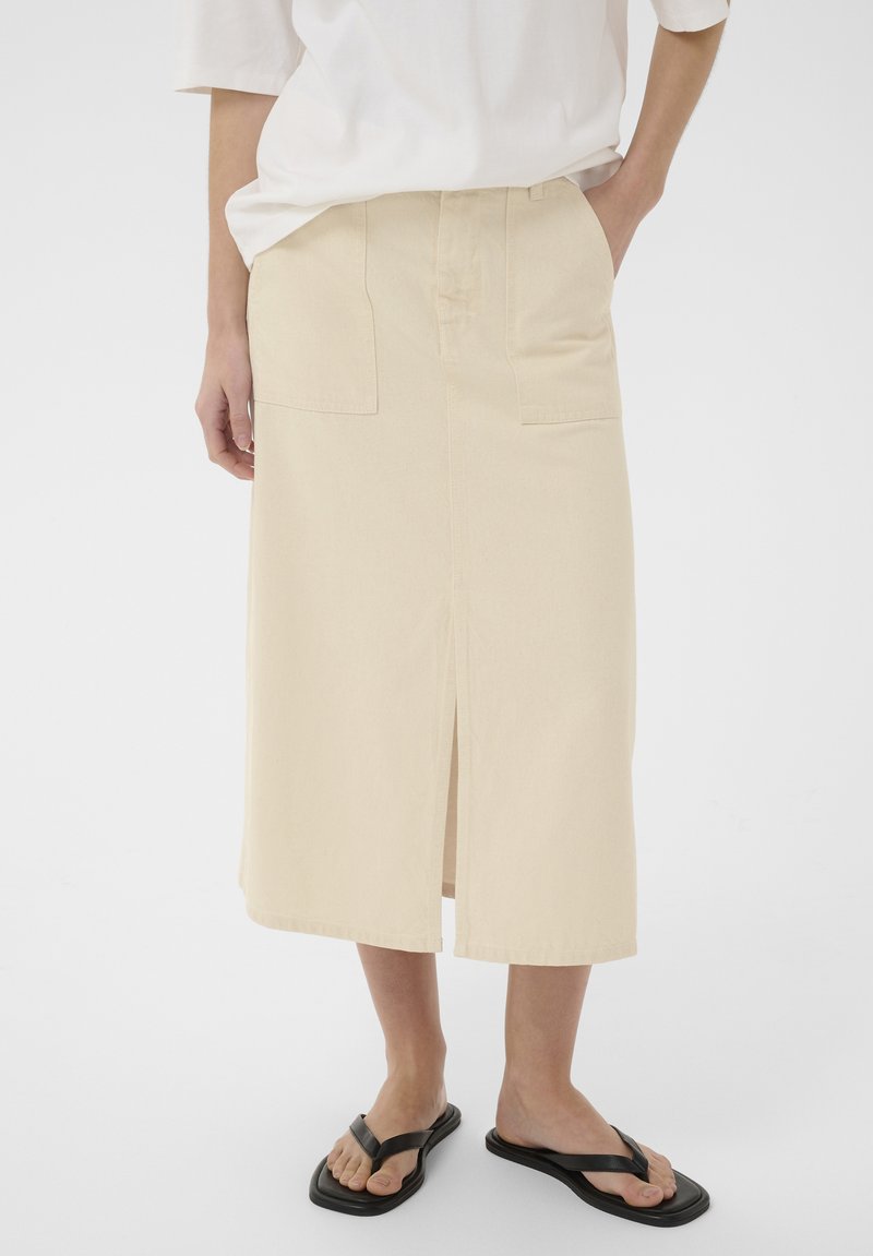 Kvinde iført en beige midi-nederdel med frontslids og store frontlommer, kombineret med en hvid løs top og sorte sandaler med firkantet tå.