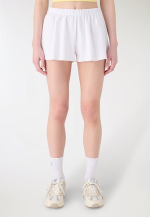 Donna con pantaloncini a vita elasticizzata bianchi, calzini bianchi alla caviglia e sneaker beige, in piedi davanti a uno sfondo bianco uniforme.