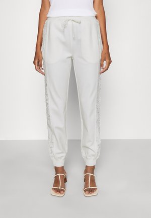 Femme portant un pantalon de jogging blanc à cordon avec des panneaux en dentelle sur les côtés et des sandales blanches à talons et lanières, debout devant un fond uni.