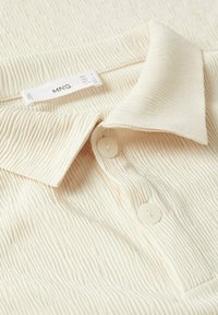Camisa de tela texturizada color crema con collar y dos botones, mostrando una etiqueta de ropa visible con detalles de marca y talla.