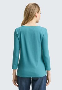 Türkises Langarmshirt mit rundem Halsausschnitt, glattem Baumwollstoff und lässiger Passform. Getragen mit dunklen Jeans, von hinten betrachtet.