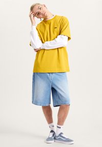 Overmaatse gele katoenen t-shirt, gelaagd met een witte lange mouwen shirt, gecombineerd met denim shorts en blauwe Vans sneakers. Accessoires omvatten ringen.