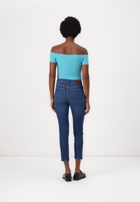 Top turquesa sin mangas combinado con jeans ajustados azul oscuro, con un parche marrón en la cinturilla y diseño clásico de cinco bolsillos.
