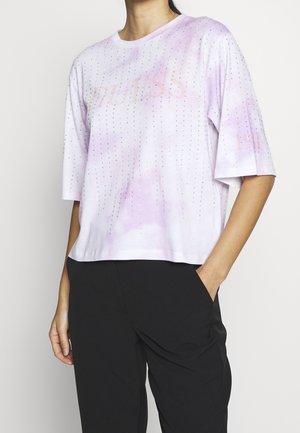 Persoon draagt een loszittend, lichtpaars tie-dye T-shirt met kleine strassteentjes en een vaag "GUESS"-tekstje, gecombineerd met een zwarte broek.