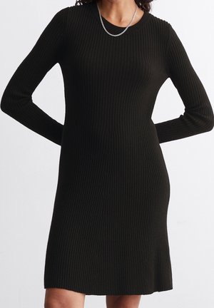 Woman wearing a long-sleeve, black ribbed knit dress with a round neckline and a silver chain necklace, standing with hands behind her back.