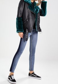 Svart läder motorcykeljacka med turkosa fuskpälskragar och manschetter, matchad med blå smala jeans med mörka sidopaneler och svarta sneakers.