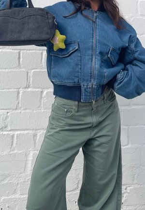 Person trägt eine kurze blaue Jeansjacke und grüne, hochtaillierte, weit geschnittene Hosen und hält eine schwarze Tasche mit einem gelben, sternförmigen Schlüsselanhänger.