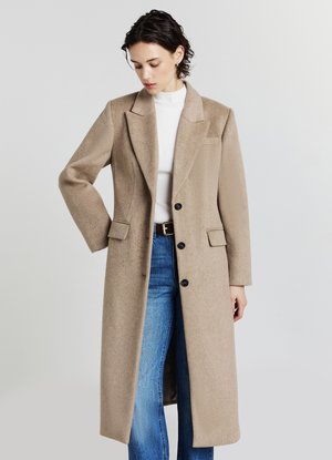 Langer beiger Mantel aus strukturiertem Stoff, mit Reverskragen, langen Ärmeln, zwei Taschen und schwarzen Knöpfen. Getragen über blauen Jeans.