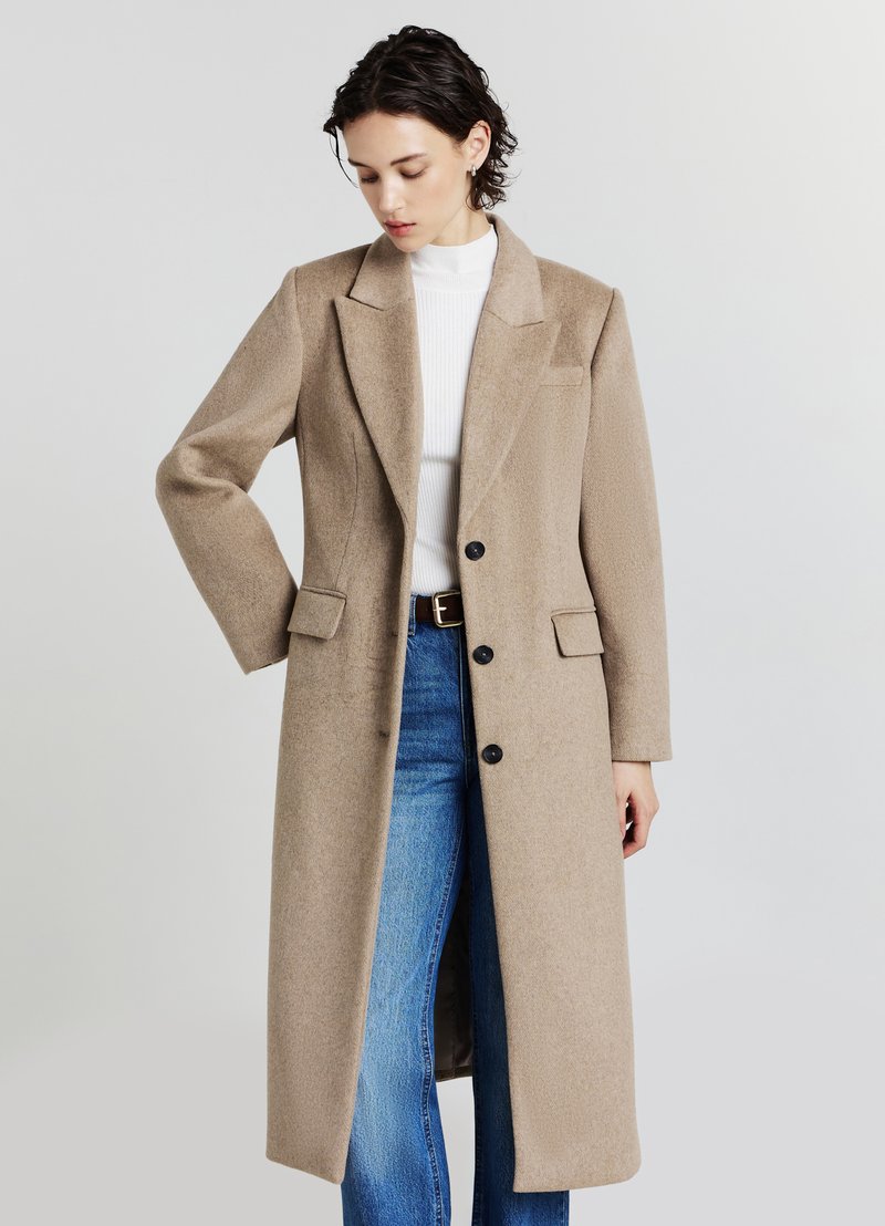 Lungo cappotto beige realizzato in tessuto strutturato, con collo a rever, maniche lunghe, due tasche e bottoni neri. Indossato sopra jeans blu.